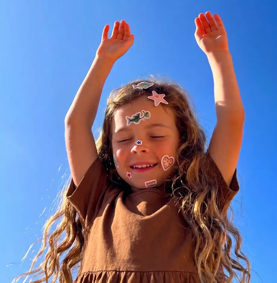 A smiling girl with stickers on her face raising her arms under a bright blue sky