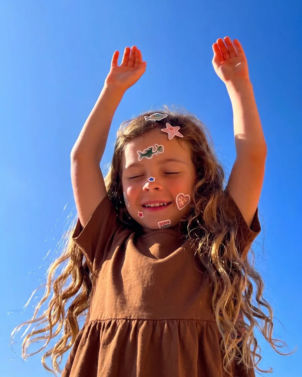 A smiling girl with long curly hair and stickers on her face, arms raised under a blue sky