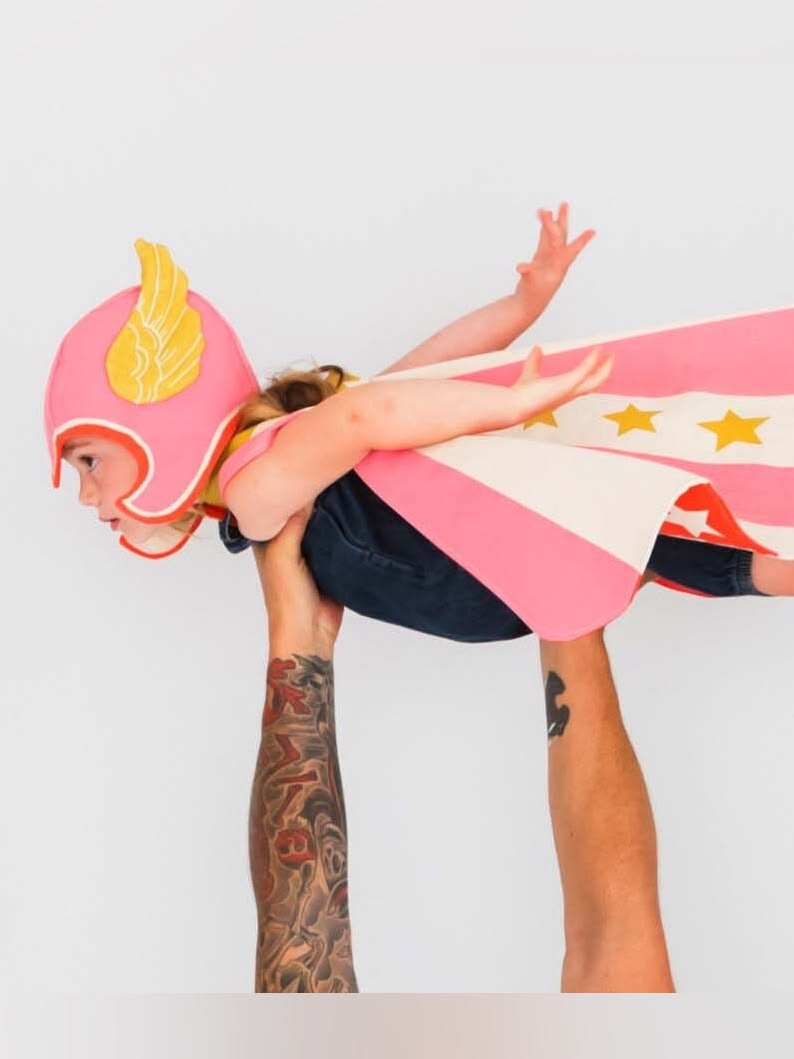 A child in a winged helmet and star cape flying in a grown-up’s hands